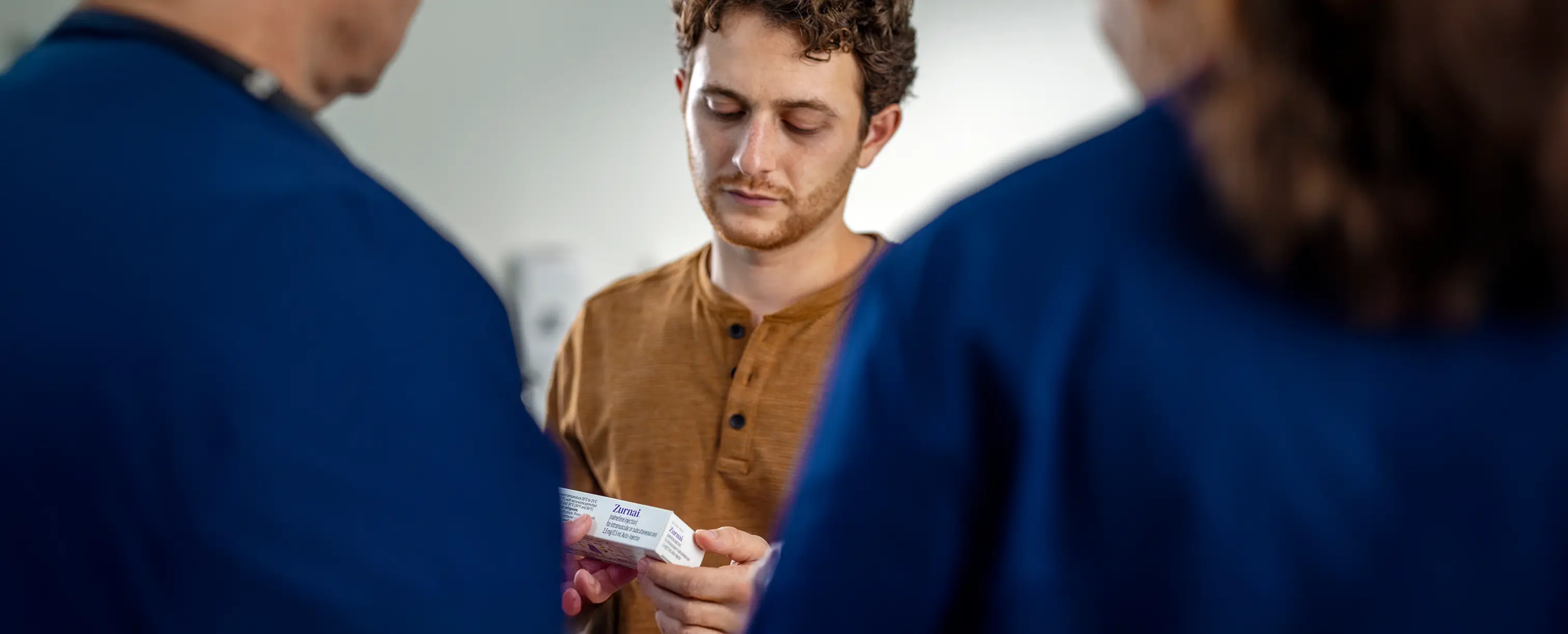 Man with brown facial hair and dark hair in a brown shirt holding the ZURNAI carton while standing before two medical professionals